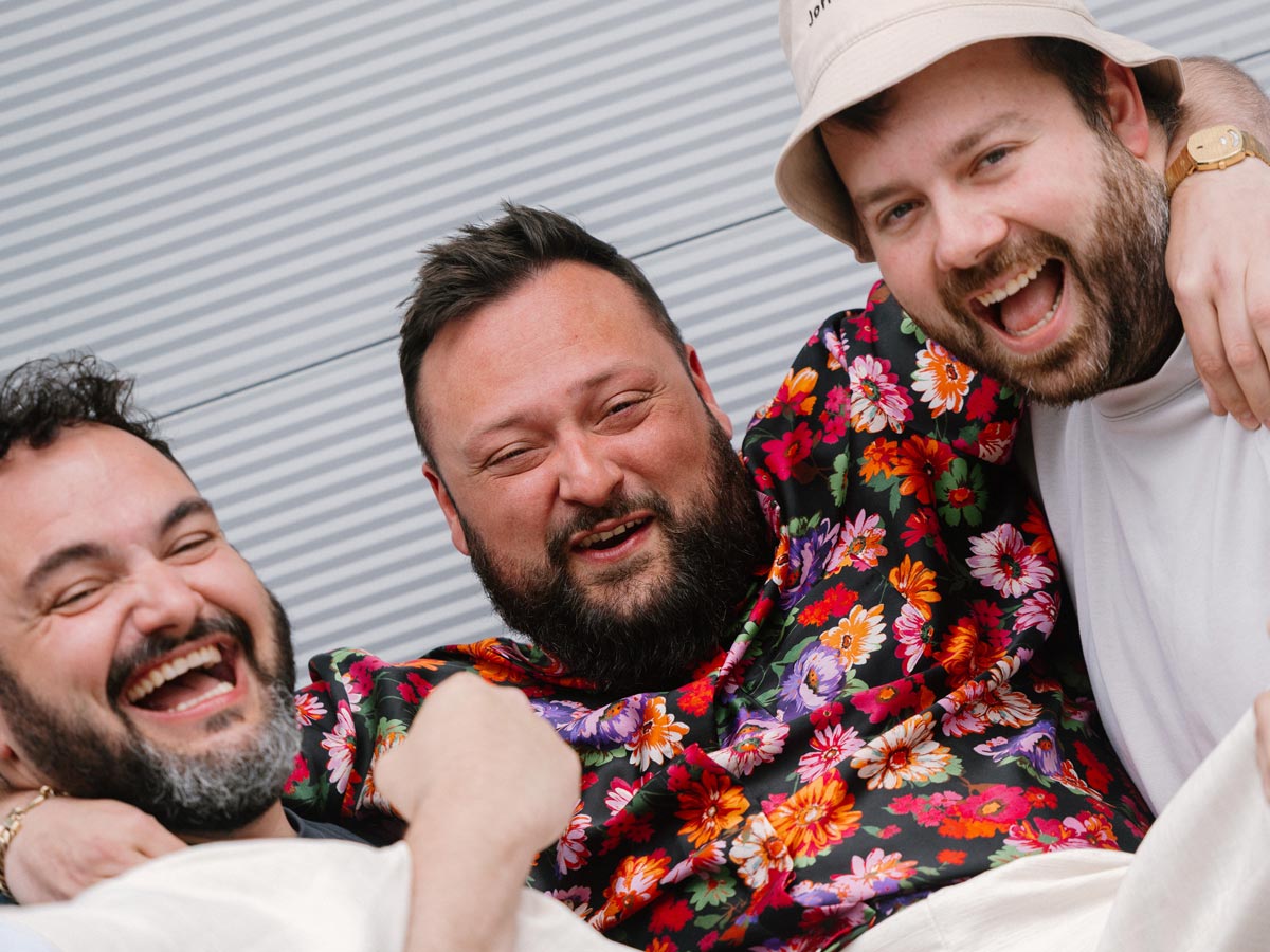 Zwei Männer heben den Mann in der Mitte nach oben. Alle lachen in die Kamera