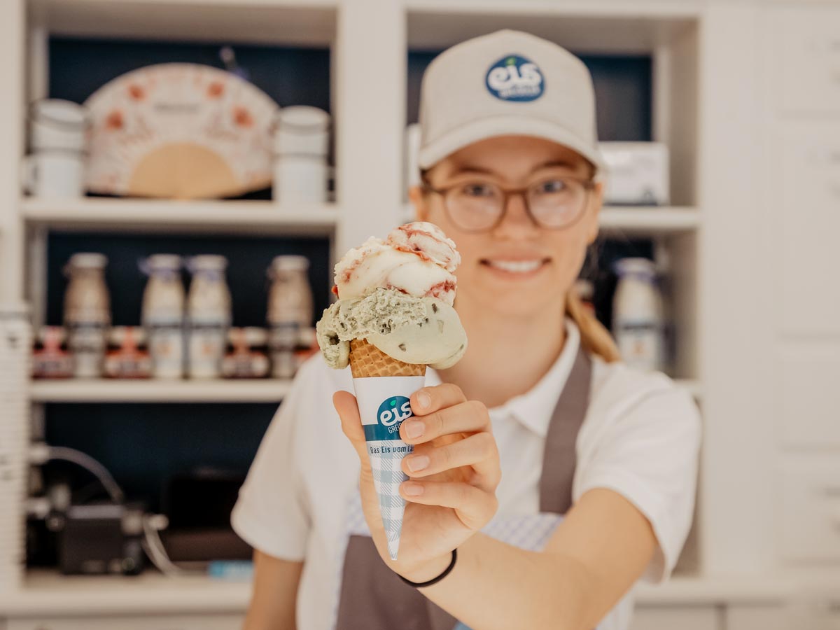 Frau hält zwei bunte Eiskugeln auf einer Waffel lächelnd in die Kamera