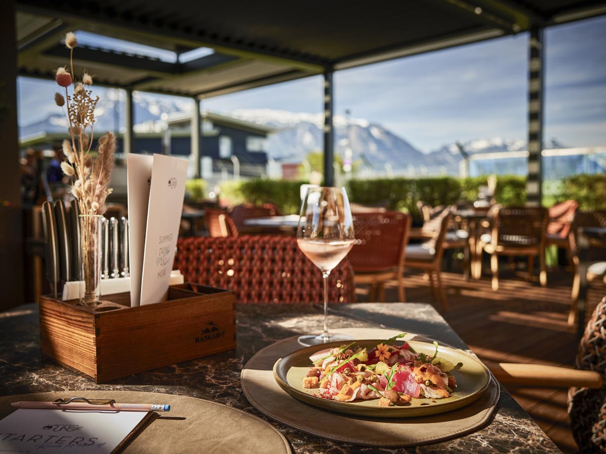 Ein Teller mit Essen und Weinglas. Im Hintergrund sind Berge zu sehen