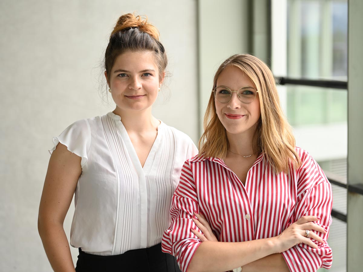 Zwei Frauen lächeln in die Kamera. Die auf der rechten Seite hat eine rot-weiß gestreifte Bluse an und die auf der linken Seite eine weiße Bluse mit ein paar Streifen